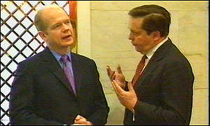 A picture of William Hague and Lord Salisbury, who at the time served under his title of Viscount Cranborne talking. On the left Hague is wering a black suit with purple tie and blue shirt, clean shaven, bald with hair on the sides. To the right is Lord Cranborne, wearing a black suit, white shirt and red tie. He is also clean shaven with combed dark brown hair. Cranborne is gesturing with both hands to Hague in conversation.