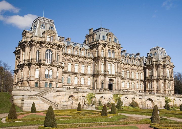 A landscape picture of Bowes Museum in Barnard Castle. In the foreground at the bottom is a smartly shaped garden with a winding path. In the background is blue sky. In the middle is Bowes Museum, a large light brown stone building with four distinct windowed levels, with three mansard roofs on the middle, left and right protruding sections of the building.
