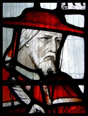Stained glass. The shoulders and head of a white man (John Morton) who has white hair and a white beard and moustache. He is wearing a red hat and red robes. He looks solemn. 