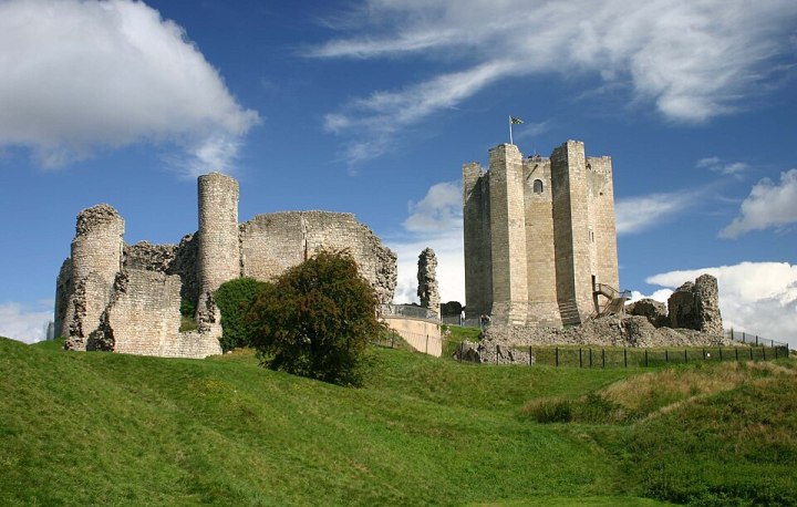 A coloured picture of the remains of Conisbrough Castle. On top of a small grassy knoll sits the castle of a light beige stonework. To the left is a ruined part of the castle, with jagged stoney edge to the remaining walls. To the right stands stands a small tower almost completely intact, with six protruding columns. The boundary wall of the castle is ruins, and instead a lower metal fence is there in its place.