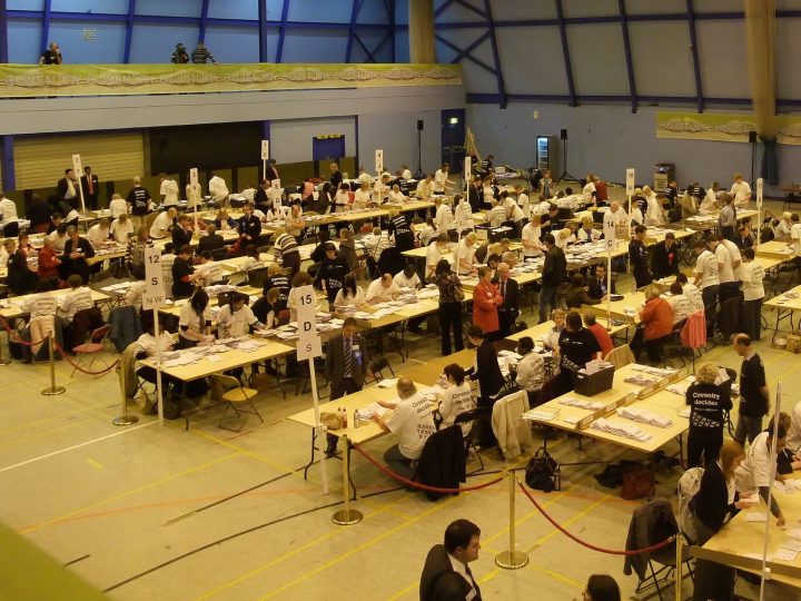 Photograph of a poll count. A number of trestle tables are set up in rows in the sports hall, with people sat in white or black T shirts sat at the tables. Large amounts of papers are in front of them, some in piles.