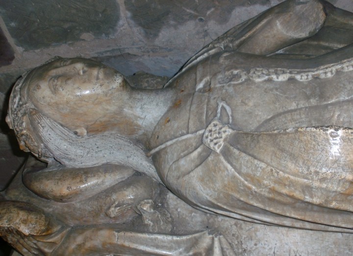 A photograph of the tomb of Elizabeth Talbot. It is a stone carving of a women lying doww. Only pictured from the waist up, she has a wreath on her head with long straight hair. She is sculpted with a dress which has roses lining it as buttons, with a cape over her shoulder.
