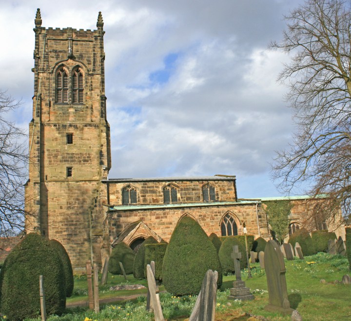 Colour photograph of St Bartholomew's Church, Elvaston, Derbyshire. In the foreground is a church yard with small landscaped bushes and various gravestones. Behind them is a Medieval style Church, with chancery and tower.