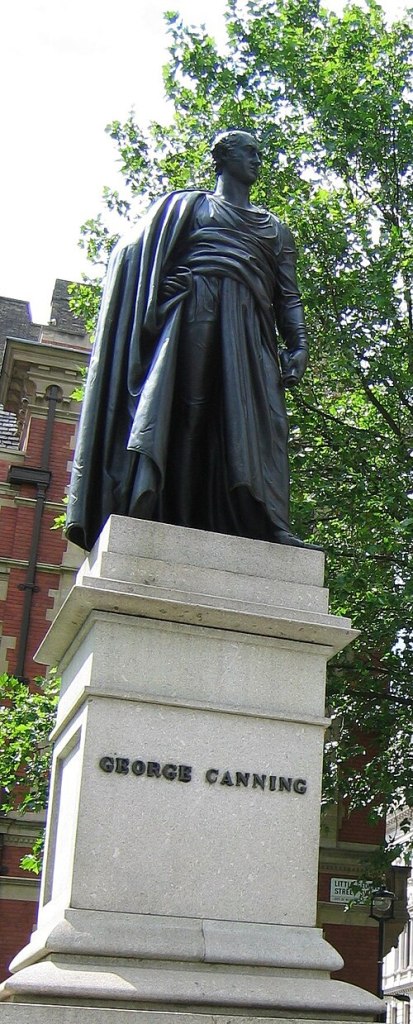 A coloured photograph of George Canning in Parliament Square. On top of a stone plinth with his name in dark capital letters, Canning stands in Roman style robes look off to his left, clean shaven with a bald patch through the middle and hair on the sides.