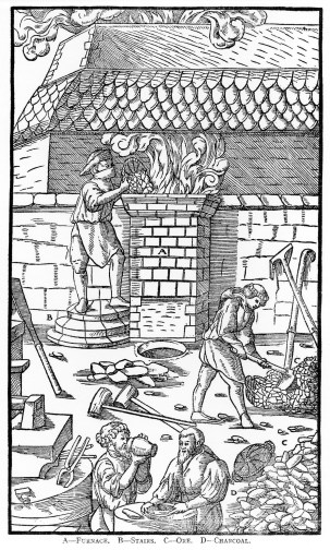 Black and white. There is an iron furnace in the background with a fire pit in the shape of a column that smoke is billowing from. There is a man stood next to this put on steps pouring in charcoal. He is wearing a hat and a mask. There is a man in front that is collecting the charcoal. At the very front of the image are two men having a drink and some food. There are tools strewn about on the floor and leant against walls.