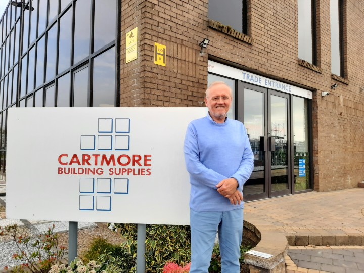 Image of Gordon Banks. He is wearing a light blue jumper and trousers, and is smiling with this hands clasped together. He is stood in front of a large building and a sign that reads 'Cartmore Building Supplies'.