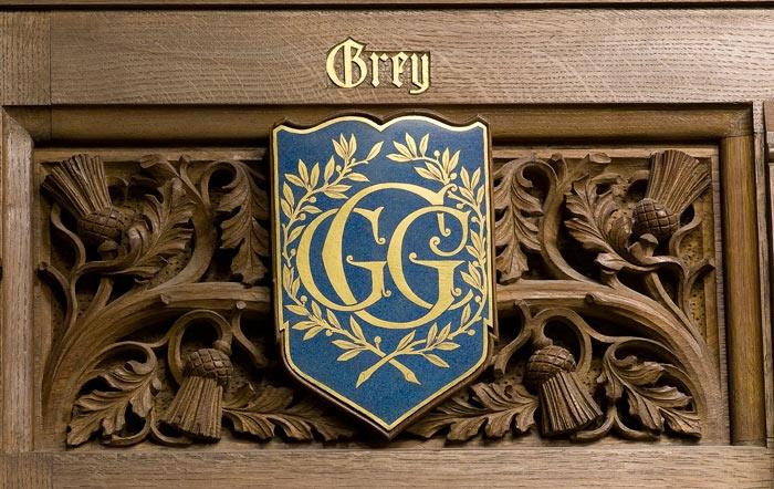 A wooden carved panel with a design of leaves and thistles. In front is a blue shield, decorated with large gold letters GCG and a laurel wreath. Above the shield in golden letters is the word Grey.