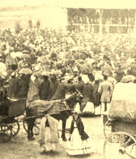 Faded sepia toned image of a large crowd. Two women stand in the foreground, in front of a horse and cart.
