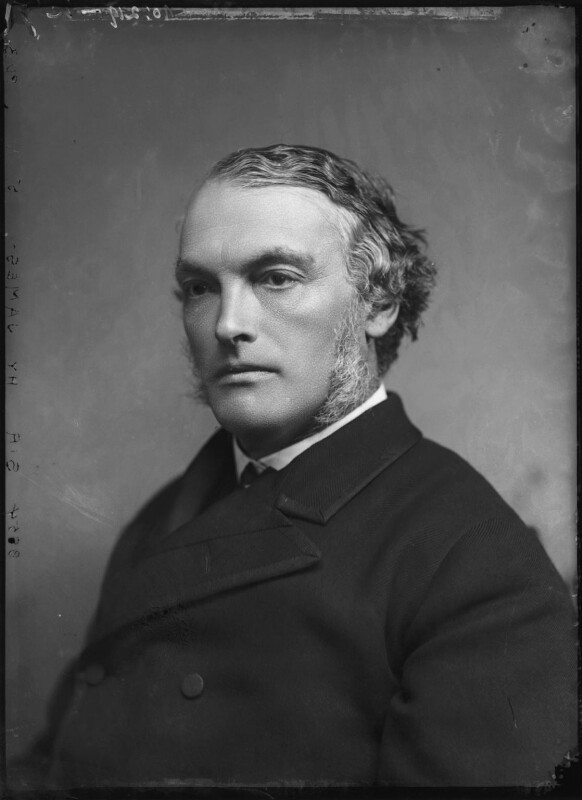 A black and white photograph portrait of a man, sitting in front of a light grey background. Sitting side on, he is wearing a double breasted black suit jacket, with a white shirt and black tie. His hair is side swept to the right and he also has long sideburns.