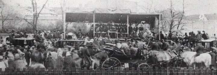 Black and white photograph showing a crowd scene. It includes horses and carriages, and at the back of the scene there is a raised platform with several men on it wearing top hats.