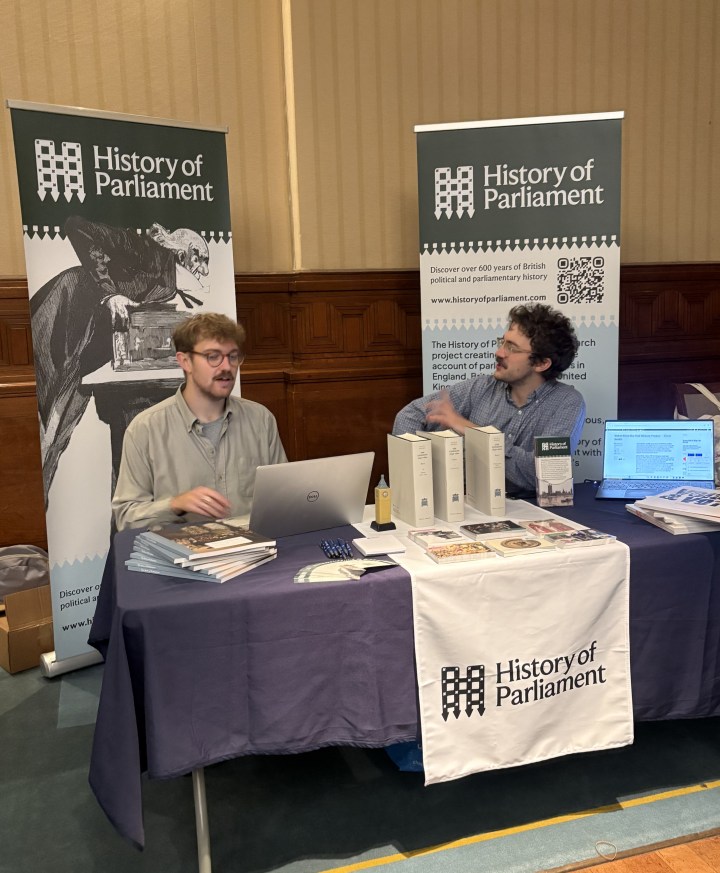 Two men are seated at a table mid conversation. The table is covered in a blue and white table cloth with History of Parliament printed on the front. Books and laptops are on the table. Two banners are behind the men, these also read 'History of Parliament'.