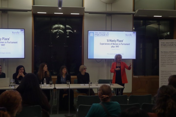 Five women are seated at a long table, looking towards a sixth who is stood at the end. She wears an orange jacket and is gesturing to a crowd of people seated in front of her. Behind the panel of people are two screens which display the words 'A Manly Place: Experiences of Women in Parliament after 1997'.