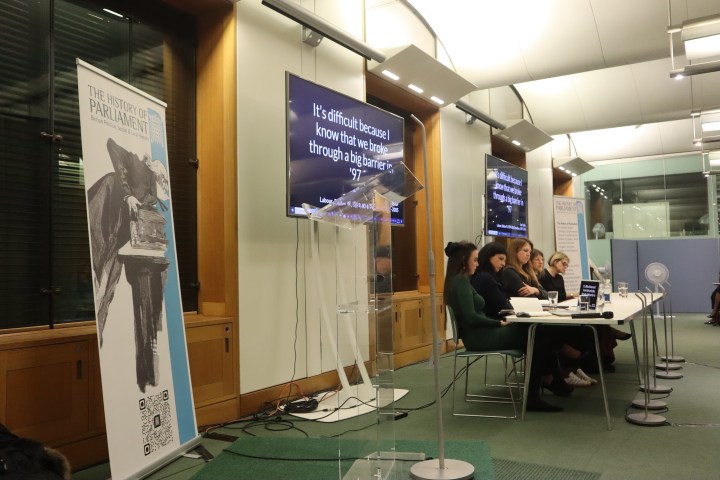 Five women sit at a long table facing the same direction. Behind them are two screens which display the words 'It's difficult because I know that we broke through a big barrier in '97'. Close to the foreground is a banner displaying the History of Parliament logo.