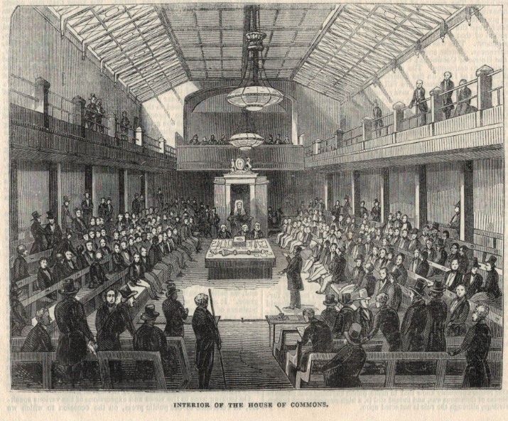 A black and white sketch of the House of Commons chamber. It is a large open space, with wooden benches for seating, many of which are occupied by men wearing suits. There are also seating galleries on either side on the upper level. It is a plainly decorated room. Two large lights hang down from the ceiling. The Speaker of the Commons is sitting in his chair, with three clerks sitting at the desk in front of him. Above his chair there is the reporters' gallery where a number of men are sitting.