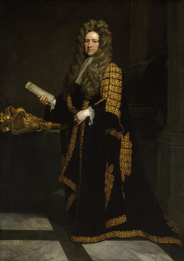 A full-length portrait of John Smith, Speaker of the House of Commons. He is standing in his full Speaker robes, which are black with heavy gold detailing. He is clean shaven with a long grey curly wig. He is holding a rolled up parchment in his right hand. Behind him is the golden Speaker's mace laying on a table to his left, and the Speaker's chair faintly to his right, with two fluted pillars either side of the chair. 