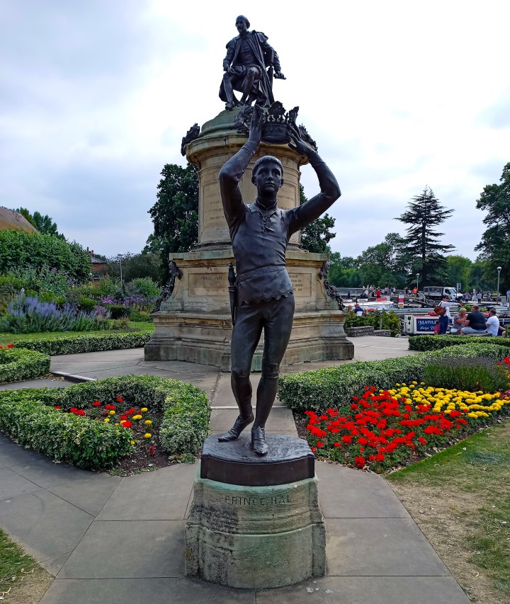 A statue of a boy holding a crown with a larger statue in the background of a man siting on a seat