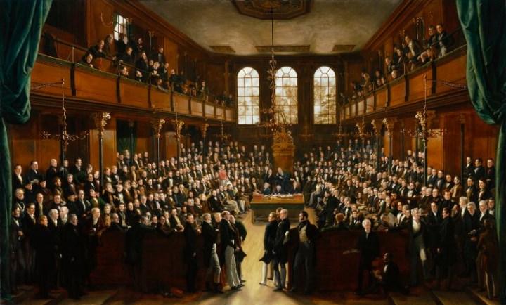 A coloured painting of the House of Commons chamber before the fire of 1834. looking into the chamber, in the middle at the back is the ornate golden speakers chair, with the Table of the House in front of it, each side, the chamber is full of MPs sitting on four rows of benches either side, there are also people standing behind and around the side. Above each side is a viewing gallery also full of people overlooking the chamber. The chamber walls a wooden with a dark golden brown polish, with black and gold detailed post holding up the galleries. There is a golden chandelier hanging low over the Table of the House, and behind the speaker is a set of three tall slim windows. 