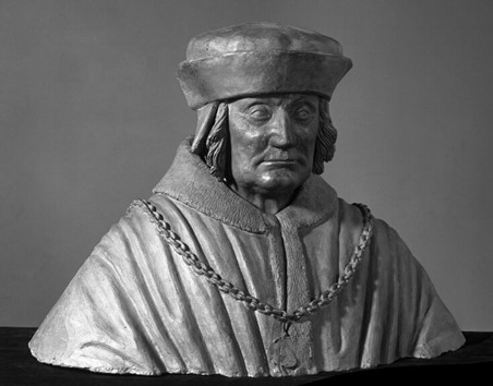A photograph of a grey bust of Sir Gilbert Talbot from the chest up, shot against a slightly darker grey background. The man is wearing a robe with a chain across the front. He has long hair, just past his head, and is wearing a flat hat with a large rim folded upwards.