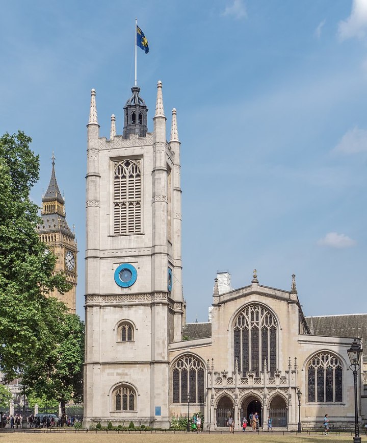 An image of a church, St Margaret's Westminster. The building is is from the centre to the right of the image. TO the left of the building is a large church tower, with a blue and yellow flag at the top. Halfway up there are two blue dials, one on each visible side of the tower. To the right the building is considerably smaller, with the entryway into the church and three larger ornate windows. To the left in the background you can see Big Ben.