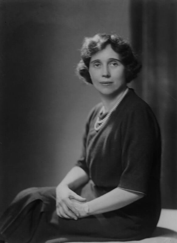 Half length black and white photograph of a young woman. She is seated with her hands clasped on her lap. She is wearing a black dress with elbow length sleeves and a double string pearl necklace. She has short dark wavy hair.