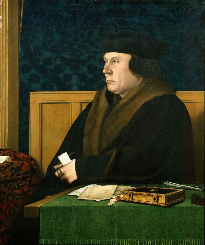 Painted half portrait of Thomas Cromwell. He is sat at a table covered with a green tablecloth, and in front of a wall covered in blue flocked wallpaper and wooden panelling. He is wearing a flat black hat and a brown coat with brown fur collar trim. He has pale skin. His left hand is leaning on the table, holding a piece of paper, and next to a book, quill and various other letters. 